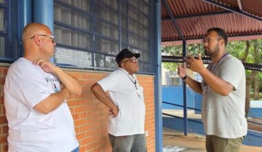 A imagem mostra três homens conversando em pé do lado de fora de um prédio. O homem mais à esquerda é careca, usa óculos escuros, uma camiseta branca e shorts jeans. Ele está encostado em uma parede de tijolos vermelhos. O homem do meio usa um boné preto, óculos, uma camiseta branca e calças jeans cinzas. Ele tem uma mão apoiada na cintura. O homem mais à direita usa óculos, uma camiseta cinza e calças verdes. Ele está gesticulando com as mãos enquanto fala com o homem do meio. Há uma cadeira branca e uma garrafa de água azul no chão atrás dos dois homens mais à direita. O prédio atrás deles tem janelas grades e um telhado de metal cor de ferrugem. O chão é de concreto rachado.