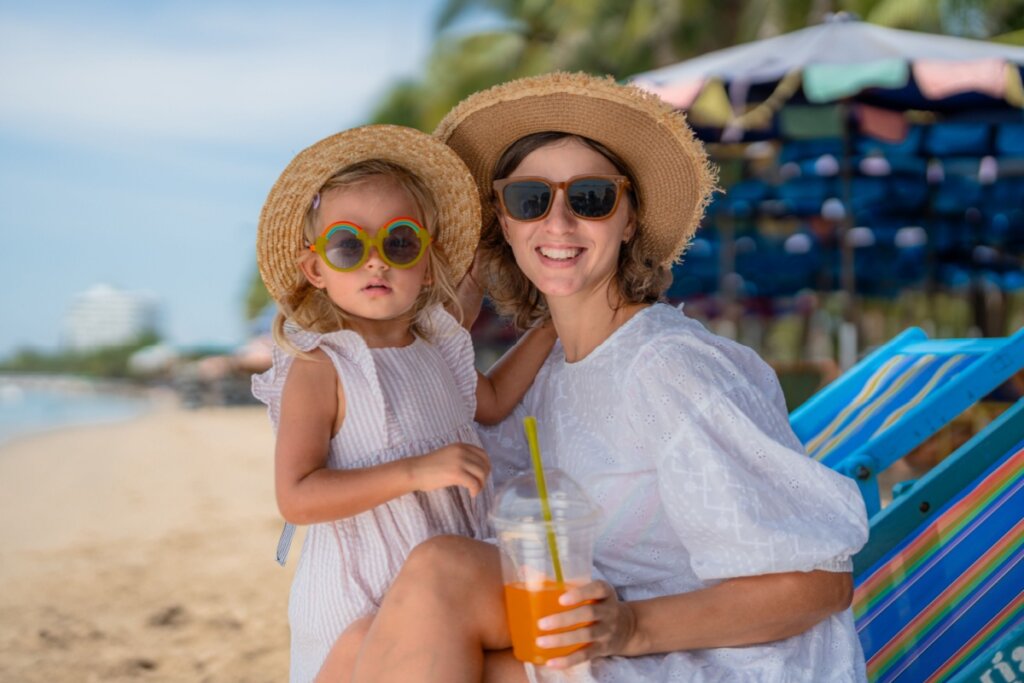 Mãe com filha no colo, ambas usando roupas brancas, chapéu e óculos de sol 