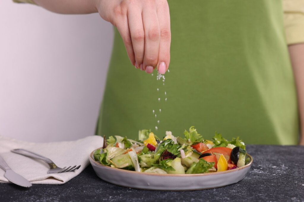 Mão feminina colocando sal em uma salada com folhas e frutas em um prato cinza 
