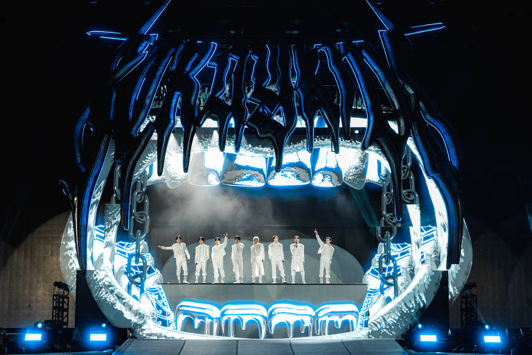 A imagem mostra um palco de show com uma estrutura grande e futurista, que se assemelha a uma boca aberta. Sete artistas vestidos de branco estão em pé no centro do palco, com luzes azuis e brancas iluminando a cena. O fundo é escuro, destacando a iluminação do palco.