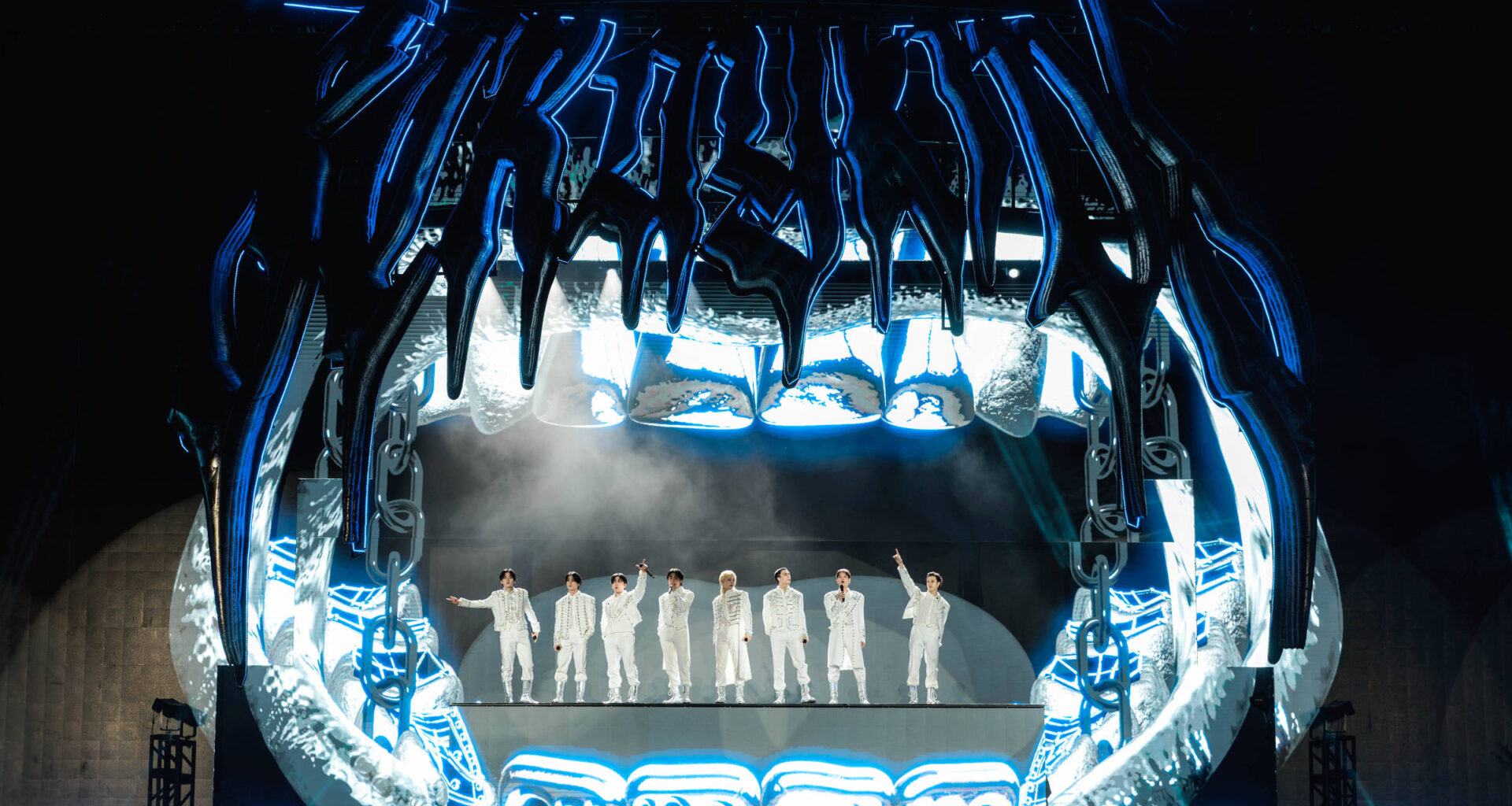 A imagem mostra um palco de show com uma estrutura grande e futurista, que se assemelha a uma boca aberta. Sete artistas vestidos de branco estão em pé no centro do palco, com luzes azuis e brancas iluminando a cena. O fundo é escuro, destacando a iluminação do palco.