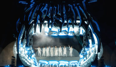 A imagem mostra um palco de show com uma estrutura grande e futurista, que se assemelha a uma boca aberta. Sete artistas vestidos de branco estão em pé no centro do palco, com luzes azuis e brancas iluminando a cena. O fundo é escuro, destacando a iluminação do palco.