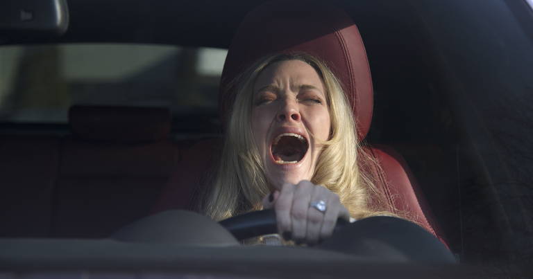 Mulher loira ao volante de carro com interior vermelho, com boca aberta em grito ou expressão de susto, segurando o volante com a mão esquerda.