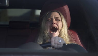 Mulher loira ao volante de carro com interior vermelho, com boca aberta em grito ou expressão de susto, segurando o volante com a mão esquerda.