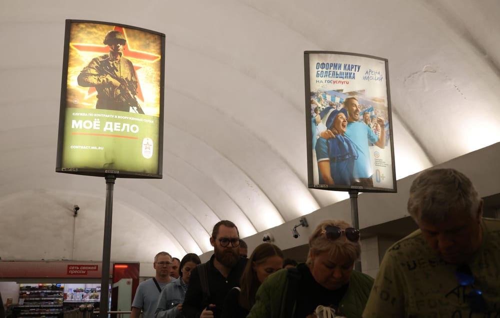 Vários passageiros passam por cartazes de propaganda do regime russo instalados na estação de metro de Dostoyevskaya, em São Petersburgo. No cartaz da esquerda é possível ver uma imagem de promoção das Forças Armadas (Contributor/Getty Images)