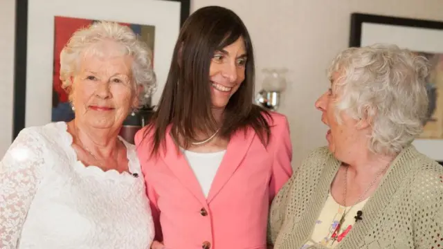 Uma mulher de jaqueta rosa está entre duas mulheres idosas, que sorriem e riem