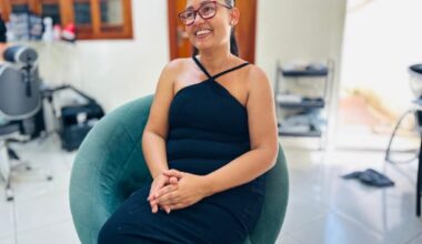 A mulher está sentada em uma cadeira azul-petróleo, vestindo um vestido preto justo com alças finas. Ela usa óculos de aro vermelho e tem o cabelo preso em um rabo de cavalo. As mãos dela estão entrelaçadas no colo. O fundo da imagem está desfocado, mas é possível ver uma janela com moldura de madeira e prateleiras com objetos.