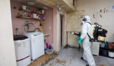 A imagem mostra uma pessoa vestindo um traje de proteção completo, incluindo um macacão branco, luvas verdes, máscara de gás e óculos de proteção, enquanto opera um pulverizador com mochila. A pessoa está em um cômodo que parece ser uma área de serviço ou lavanderia, com máquinas de lavar roupa, prateleiras com produtos de limpeza e uma pia. O chão está sujo e descascado em algumas áreas, e as paredes parecem desgastadas. Há também um varal improvisado com roupas penduradas no fundo. A pessoa está direcionando o bico do pulverizador para a parede, possivelmente realizando um processo de desinfecção ou dedetização.