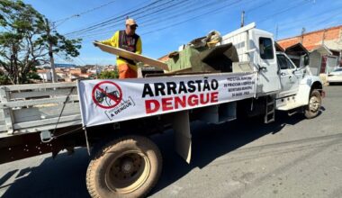 A imagem mostra um caminhão branco com uma faixa publicitária presa na lateral, que anuncia uma campanha contra a dengue. A faixa exibe um desenho de um mosquito com um sinal de proibido sobre ele, acompanhado do slogan "TODOS CONTRA A DENGUE". Abaixo, em letras maiores, lê-se "ARRASTÃO DENGUE". À direita da faixa, há um texto em letras menores que diz "PREFEITURA MUNICIPAL DE PIRACICABA A SERVIÇO DA SECRETARIA MUNICIPAL DA SAÚDE - PMCA". Um homem, vestindo um boné, óculos escuros e uma camiseta amarela com detalhes pretos e laranjas, está em cima do caminhão, segurando uma prancha de madeira. O caminhão está carregado com diversos objetos, incluindo móveis e outros materiais. A cena se passa em uma rua, com casas e árvores ao fundo, sob um céu azul claro. O logotipo "ECOTERRA" aparece duas vezes no caminhão.