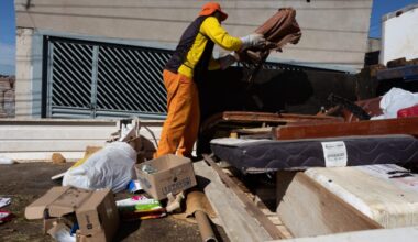 Um trabalhador com roupa laranja e amarela está a descarregar uma cama de um camião de lixo. O camião está cheio de entulho, incluindo um colchão, pedaços de madeira e outros detritos. A cena parece ser uma obra de construção ou uma limpeza de casa.