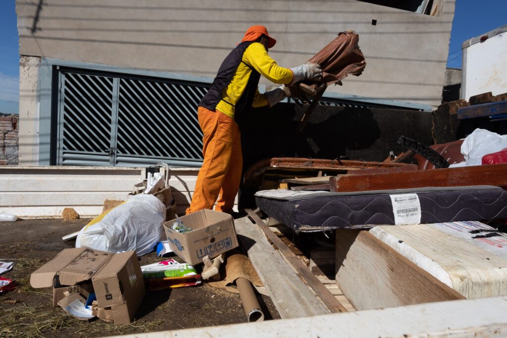 Um trabalhador com roupa laranja e amarela está a descarregar uma cama de um camião de lixo. O camião está cheio de entulho, incluindo um colchão, pedaços de madeira e outros detritos. A cena parece ser uma obra de construção ou uma limpeza de casa.