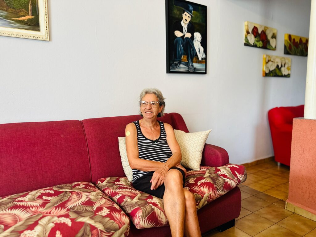 Uma mulher com cabelos grisalhos e óculos está sentada em um sofá vermelho. Ela usa um colete listrado em preto e branco e shorts pretos. Um curativo redondo e claro está visível em seu braço esquerdo. Ela sorri e olha para a direita. O sofá tem almofadas com um padrão floral vermelho e branco. Atrás dela, na parede branca, há várias pinturas. Uma delas é um retrato de Charlie Chaplin com um cachorro. Outras duas pinturas menores mostram flores vermelhas e amarelas. Há também um sofá vermelho em forma de poltrona ao fundo, à direita. O piso é de ladrilhos em tons de bege e marrom.