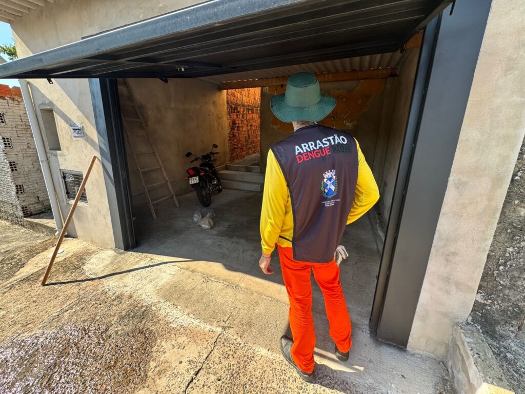 A imagem mostra uma pessoa vestindo um chapéu verde, uma camisa amarela de manga comprida e uma vestimenta preta por cima com a inscrição "ARRASTÃO DENGUE 2022". A pessoa também usa calças laranjas e sapatos pretos. A cena parece ser em uma área externa, possivelmente uma garagem ou um quintal, com uma construção ao fundo. Há uma moto estacionada, uma escada encostada na parede e materiais de construção visíveis. A iluminação sugere que é dia.