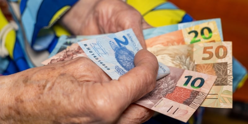 Mãos envelhecidas de uma pessoa idosa segurando notas de dinheiro para ilustrar a matéria sobre desigualdade social e envelhecimento.