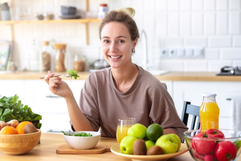 mulher de cabelo preso, blusa marrom, sorrindo, sentada e segurando garfo com salada. à frente dela, pote com salada, copo com suco e prato com frutas
