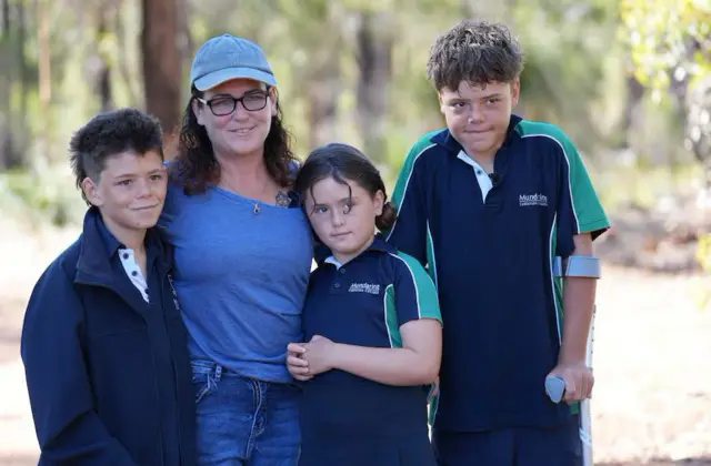 Beau está ao lado de sua mãe, que veste uma camiseta azul, e de sua irmãzinha, Grace. Todas as crianças estão de uniforme escolar. Austin está apoiado em muletas.