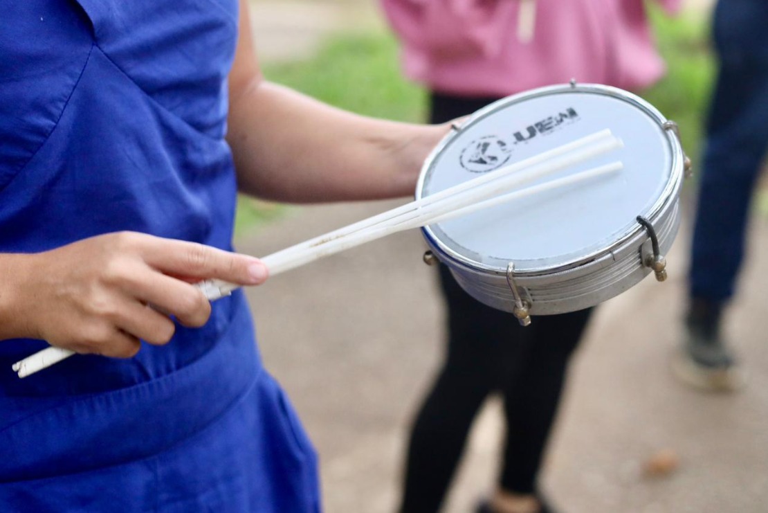 A imagem mostra, em primeiro plano, uma pessoa vestindo uma roupa azul segurando um tamborim, instrumento típico do samba, com uma das mãos, enquanto na outra segura duas baquetas brancas posicionadas sobre a pele do instrumento. O foco está nas mãos e no tamborim, sugerindo que a pessoa está prestes a tocar ou em um momento de apresentação. É possível ver a marca do instrumento impressa na pele. Ao fundo, de forma desfocada, aparecem outras pessoas em pé, usando roupas casuais, o que indica que a cena ocorre em um ambiente ao ar livre, possivelmente em um ensaio, atividade cultural ou evento comunitário. A composição da imagem transmite a ideia de ritmo, música e participação coletiva, associada ao samba e à cultura popular brasileira, especialmente relacionada a contextos como o Carnaval ou apresentações culturais.