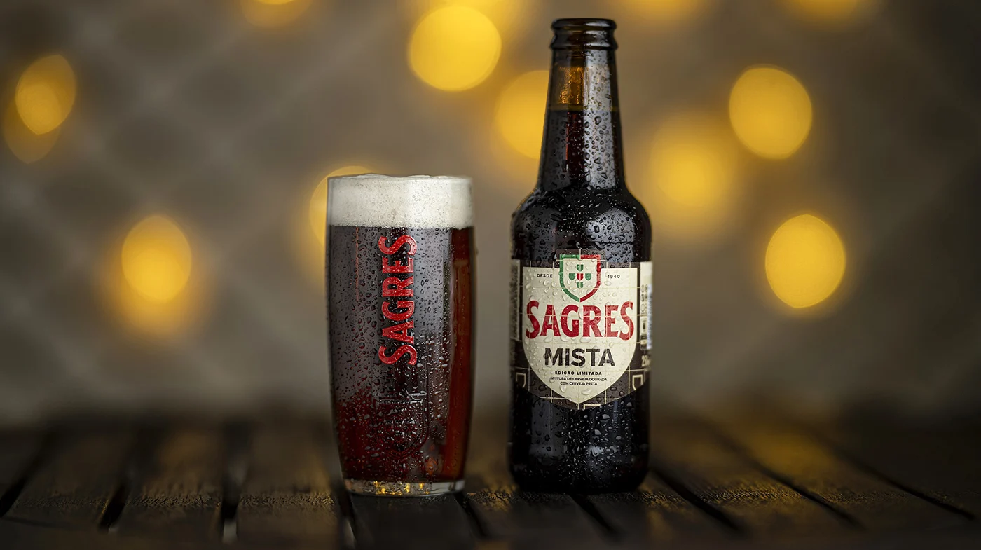 Copo e garrafa de cerveja Sagres com condensação, sobre mesa de madeira, com luzes desfocadas ao fundo