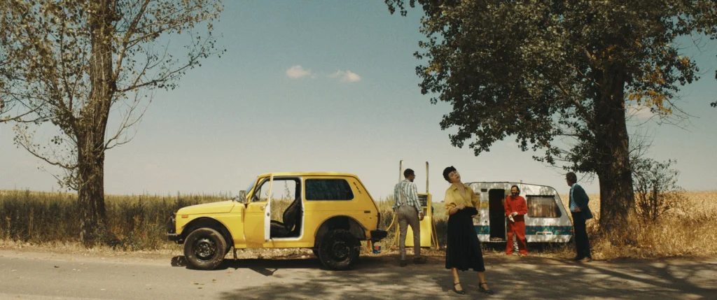 Imagine dintr-un film, o masina galbena si trei personaje, un barbat in alb, o femeie in galben si negru si una in rosu