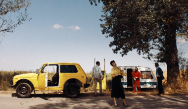 Imagine dintr-un film, o masina galbena si trei personaje, un barbat in alb, o femeie in galben si negru si una in rosu