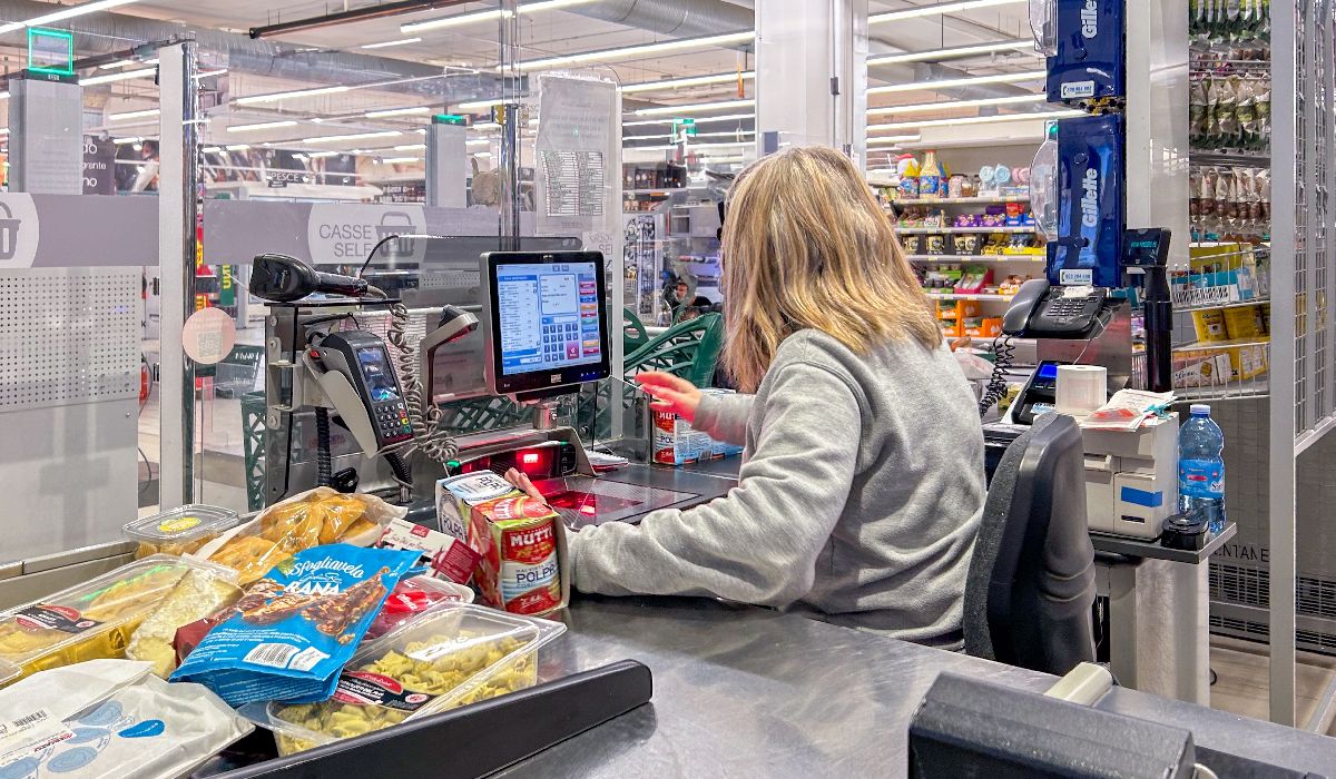 O femeie blondă îmbrăcată într-un pulover gri scanează produsele alimentare la casa de marcat a unui supermarket, înconjurată de rafturi cu produse și de un ecran digital.