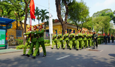 Un grup de militari vietnamezi care defilează în formație pe o stradă urbană, purtând uniforme verzi și un steag roșu.
