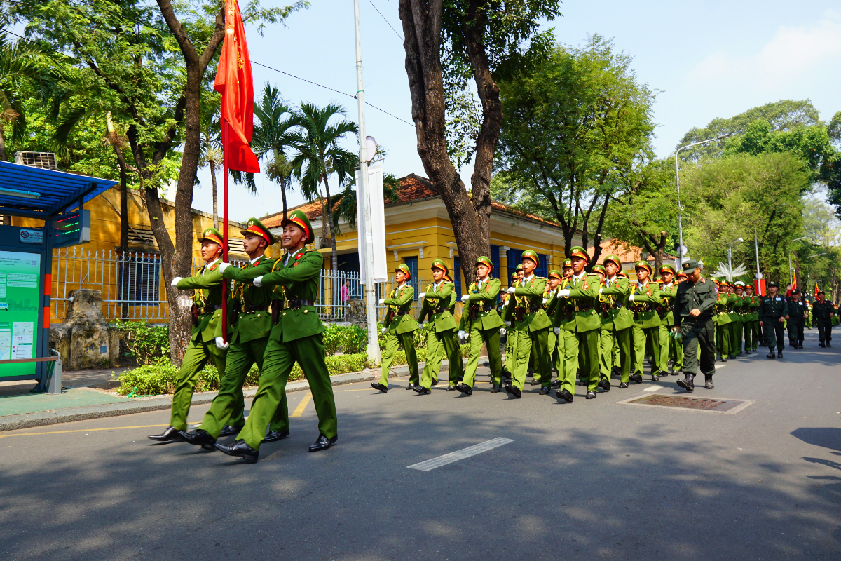 Un grup de militari vietnamezi care defilează în formație pe o stradă urbană, purtând uniforme verzi și un steag roșu.