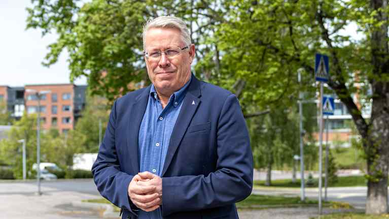 En man i blå skorta och blå kavaj står med knäppta händer och tittar in i kameran. I bakgrunden träd.