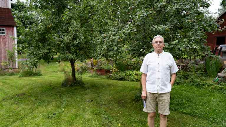 En man i vit skjorta och med beigea shorts som står vid ett äppelträd.