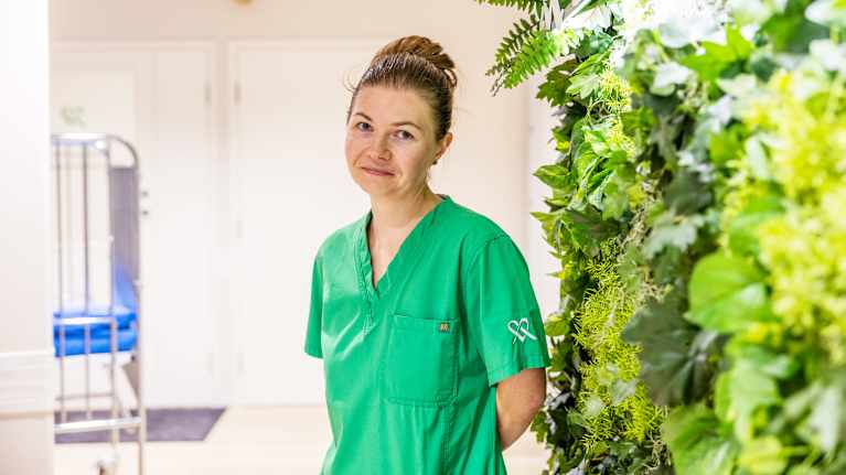 En tandläkare i gröna arbetskläder står bredvid en grön vägg av växter.