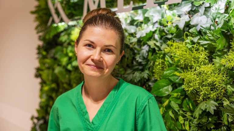 På bilden ser vi en person klädd i grön sjukvårdspersonaluniform framför en vägg dekorerad med gröna växter.