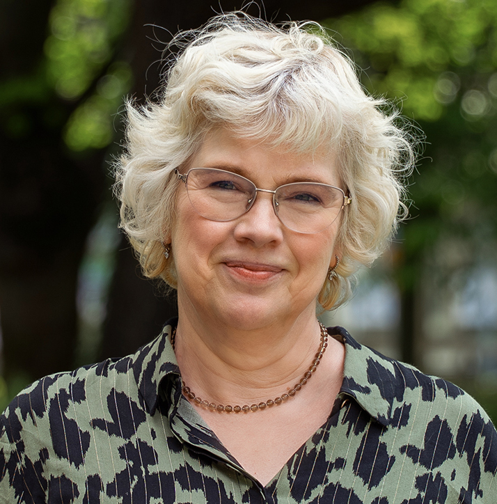 En kvinna med kort, lockigt, ljusblont hår och glasögon står utomhus. Hon har på sig en grön och svart mönstrad blus och ett pärlhalsband, med träd och grönska suddigt i bakgrunden.