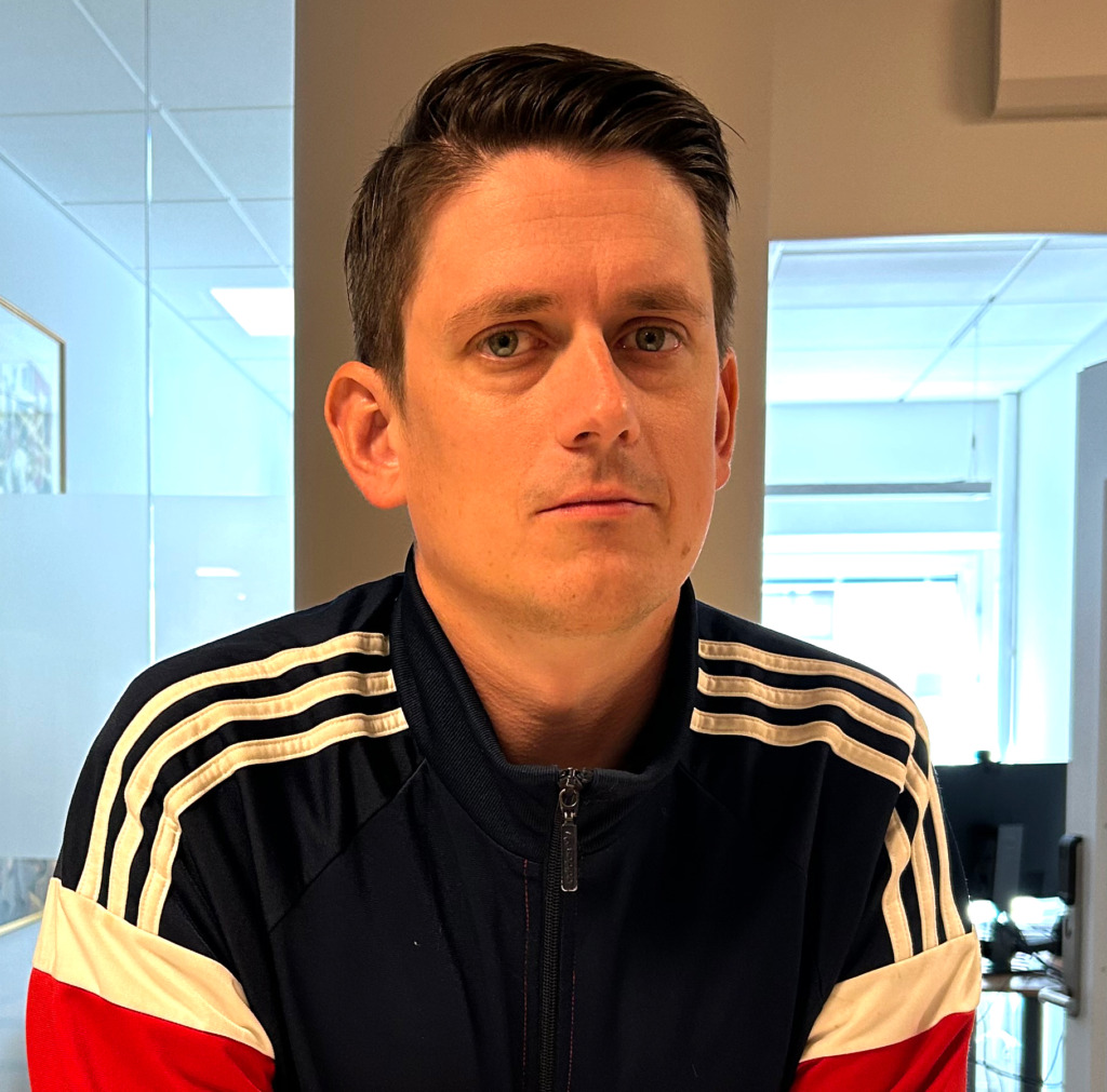 En man med kort brunt hår, klädd i en marinblå jacka med vita och röda ränder, står inomhus i en kontorsmiljö och tittar rakt in i kameran med ett neutralt ansiktsuttryck.