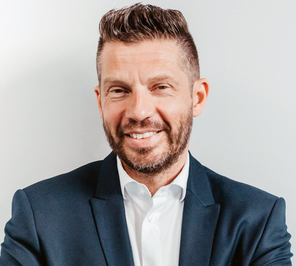 En man med kort brunt hår och vältrimmat skägg, klädd i mörk kostymjacka och vit skjorta, står framför en ljus bakgrund och ler mot kameran.