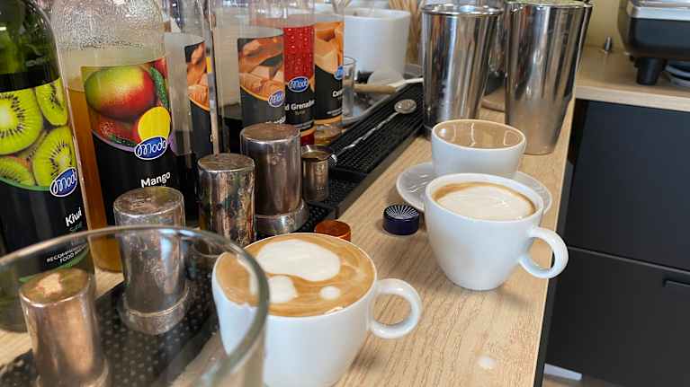 Specialkaffen på kafé: en rad flaskor med smaksirap och kryddburkar står intill tre koppar kaffe av olika slag står på en arbetsbänk i ett kafékök.