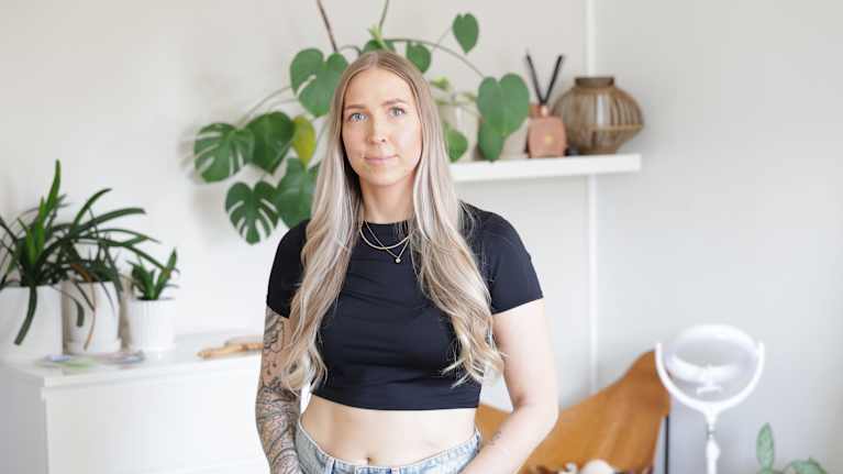 Kvinnan på bilden har långt blont hår, en svart topp och ljusa jeans. Hon befinner sig i ett rum med gröna växter i bakgrunden.