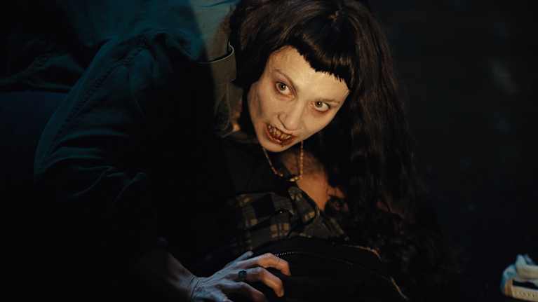 En blek kvinna med mörka kläder, blodig mun och huggtänder ligger böjd över en person som ligger på golvet.