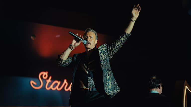 En medelålders man i svart skjorta och gråblå sidenkavaj står på en scen och sjunger i en mikrofon, den andra handen håller upp i luften.