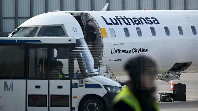 Ett Lufthansaplan står på flygplatsen med landgång och luckan till lastrummet öppna. Framför syns ett markfordon. Ett par resväskor ligger på marken..