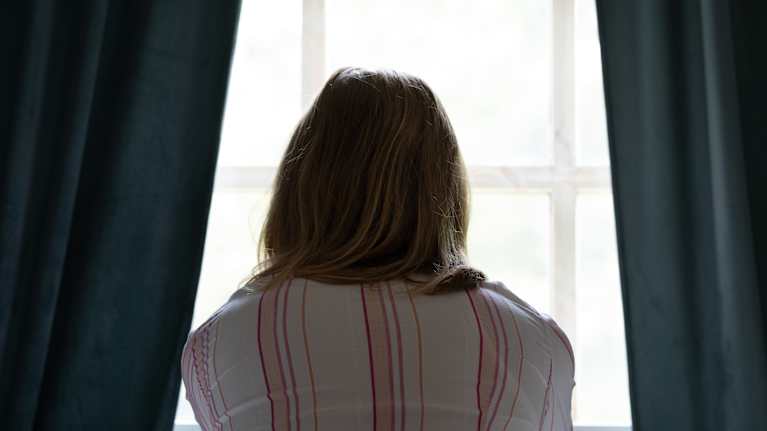 En person sitter på en säng och tittar ut genom ett fönster, med ryggen vänd mot kameran.