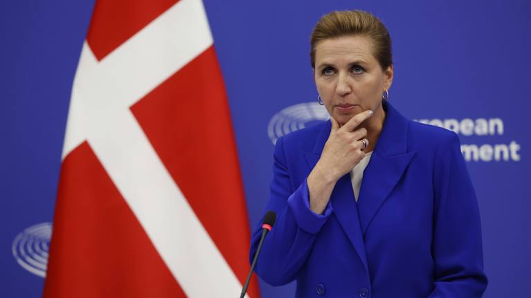 Danmarks statsminister Mette Frederiksen klädd i blått i ett talarpodium med en dansk flagga intill sig.