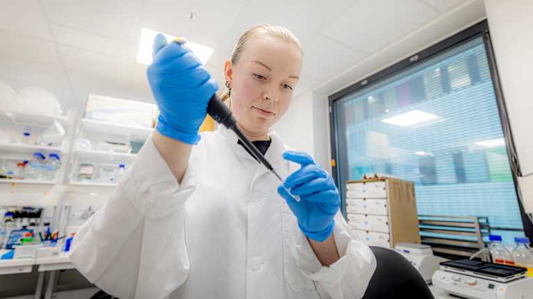 en person i en laboratoriemiljö som hanterar en pipett, iförd skyddskläder i form av en vit labbrock och blå engångshandskar. I bakgrunden syns laboratorieutrustning.