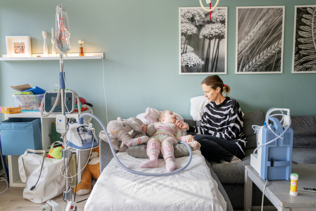 En kvinna sitter i en soffa. Ett barn ligger i en säng bredvid soffan.