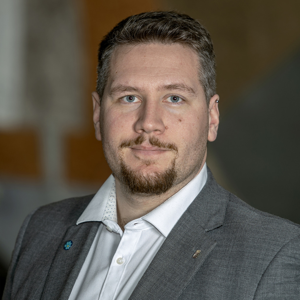 En man med kort brunt hår, vältrimmat skägg och mustasch bär en grå kostymjacka och en vit skjorta med krage och tittar in i kameran med ett neutralt uttryck. Bakgrunden är suddig.