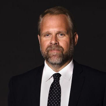 En man med ljusbrunt hår och skägg är klädd i mörk kostym, vit skjorta och svart prickig slips. Han poserar mot en enkel, mörk bakgrund och tittar direkt in i kameran med ett neutralt uttryck.