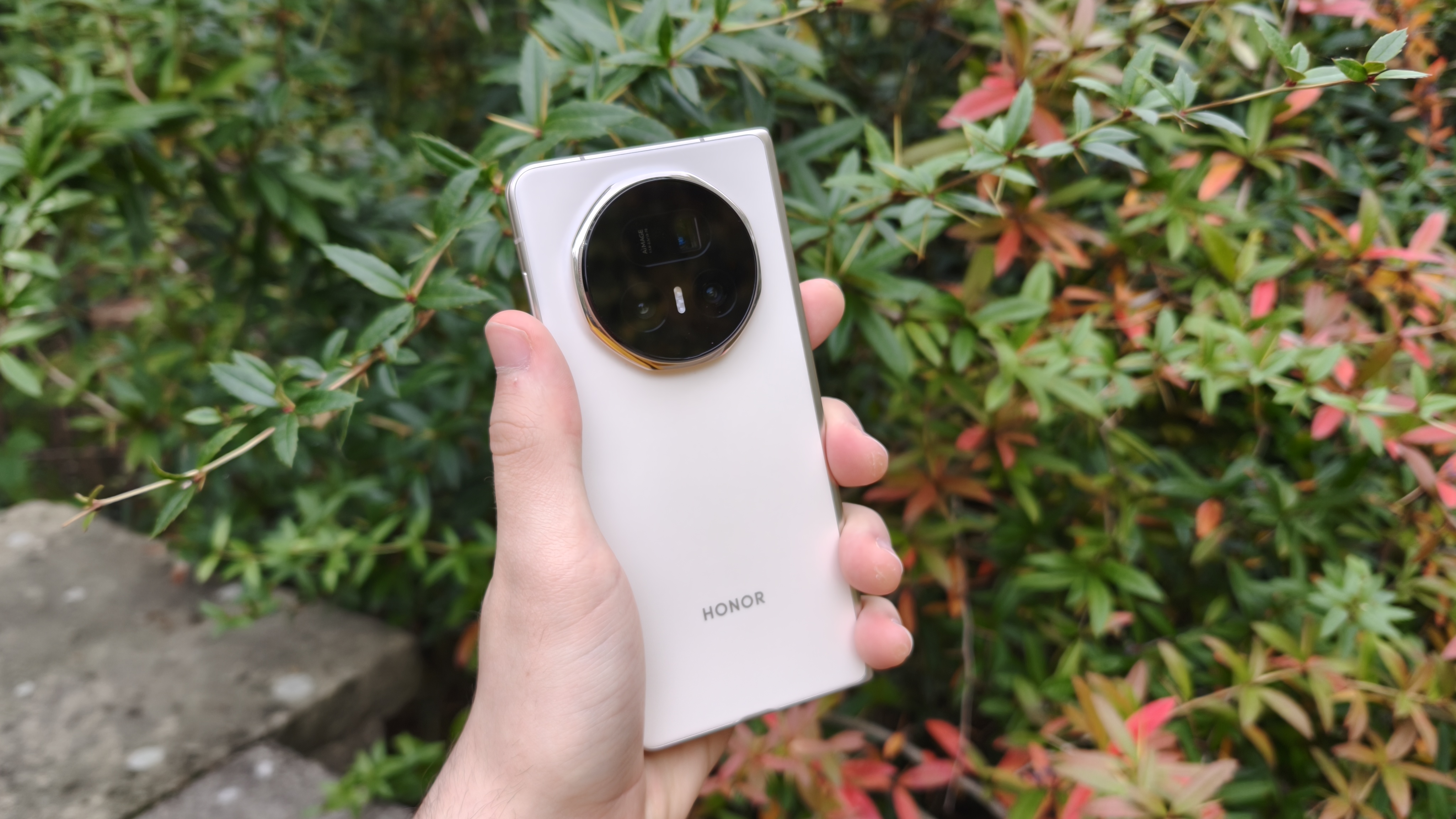 Honor Magic V5 i vitt h&aring;lls upp framf&ouml;r ett g&auml;ng buskar.