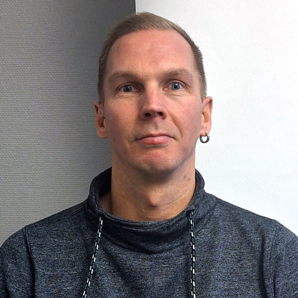 En person med kort ljusbrunt hår, klädd i en grå tröja med hög krage och ett enkelt örhänge i vänster öra, vänder sig mot kameran mot en bakgrund som till hälften är grå med textur och till hälften vit.