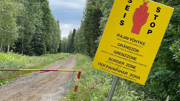 Skylt vid gränszon i skogen, med text på flera språk som uppmanar att stanna.