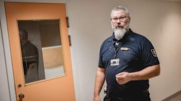 En uniformerad man står framför en orangefärgad dörr med ett fönster.