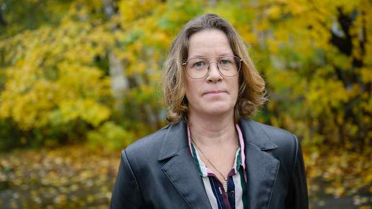 En kvinna står utomhus med en bakgrund av höstlöv i olika gula och gröna nyanser.